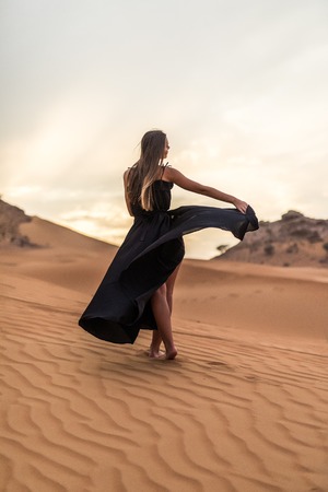 Young woman in black dress dancing in sandy desert at sunsetの写真素材