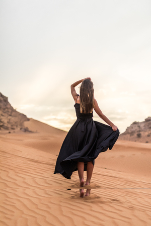Rear view of young pretty woman in black dress dancing in sandy desert at sunsetの写真素材