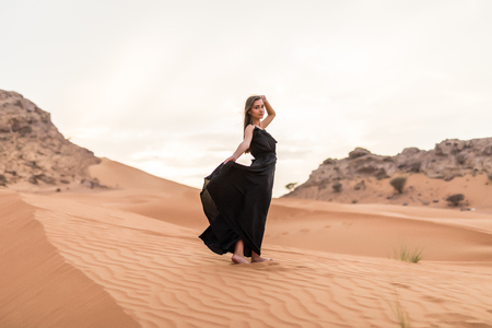 Young woman in black dress stands on dune in desert at sunny hot dayの写真素材
