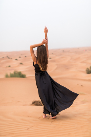 Rear view of young woman in black dress dancing in sandy desert at sunsetの写真素材