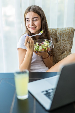 Portrait of happy young woman eating fresh salad from a bowl while sitting and using laptop computer at homeの写真素材