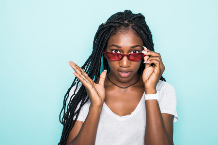 Fashion portrait of young african woman smiling looking on camera in modern sunglasses isolated over blue backgroundの写真素材