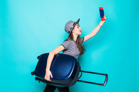 Portrait of young woman holding passport with flying tickets while standing with a suitcase isolated over blue backgroundの写真素材