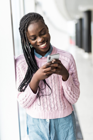 Young african woman surf in internet in mobile phone in front of panoramic windowsの写真素材