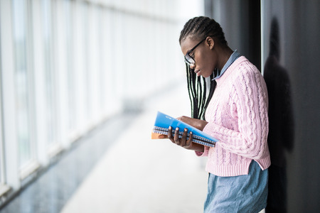Young afro american girl student in glasses holding notebooks and studying prepare for exam in modern University hallの写真素材
