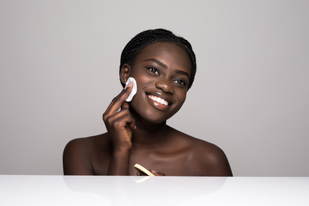 Young pretty afro american woman taking off her makeup with cotton wipe sponge isolated on whiteの写真素材