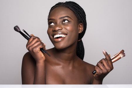 Beauty portrait of a smiling beautiful half naked african woman posing with make-up brushes and looking away over white backgroundの写真素材