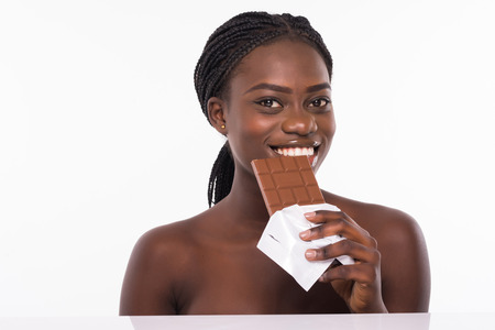 Portrait of a smiling young african woman eating chocolate bar isolated over whiteの写真素材