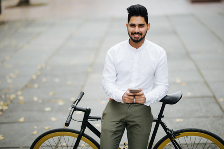 Handsome indian man in classic suit is using a smart phone and smiling while riding bicycle in cityの写真素材