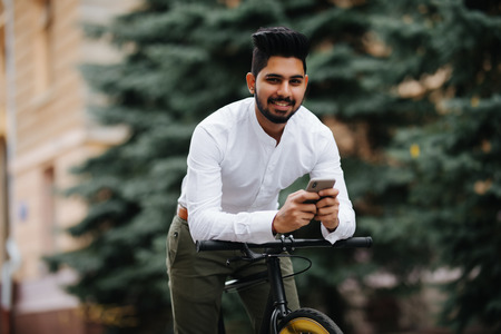 Handsome young indian man holding mobile phone while standing near his bicycleの写真素材