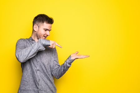 Portrait of a cheerful young man pointing fingers away at copy space on his palm over yellow backgroundの写真素材