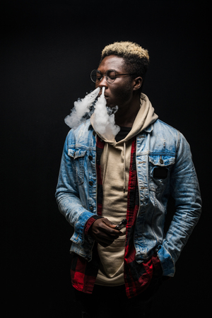 Portrait of african man it smoking an e-cigarette isolated on dark background.の写真素材