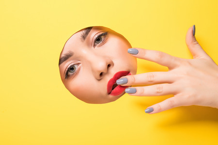 A girl with beautiful long fingers and red nails touches the edge of her eye and looks into the circular hole of the yellow paper.の写真素材