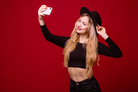 Fashion woman in black hat with blond curly hair, bright red lips taking selfie on red background. Trendy fashion styleの写真素材