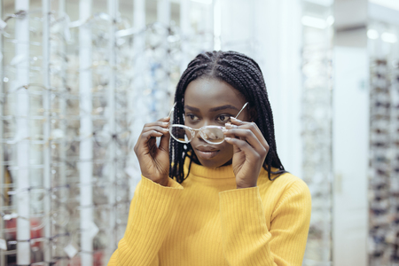 Young african pretty woman choosing eyeglasses frames in Optical Store. Cute girl deciding for glasses of different shapes and colorsの写真素材