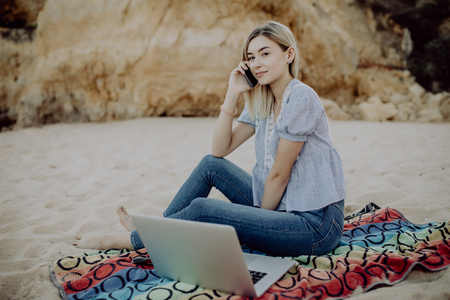 Young woman lying on white sand with laptop and talking by her phone on the rocky beachの写真素材