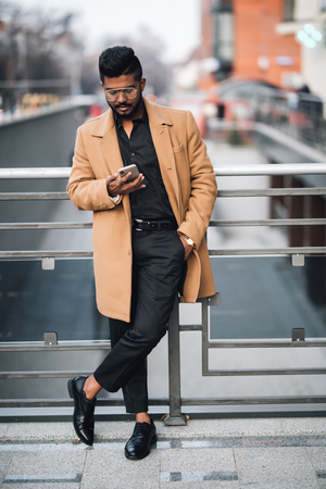 Young indian businessman in coat serfing in internet from phone in the city streetの写真素材