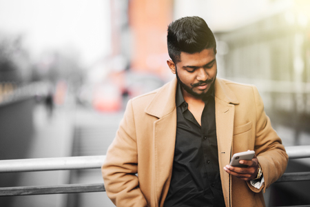 Portrait of bearded indian man in coat use internet networks from phone on the streetの写真素材