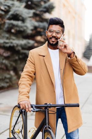 Handsome young indian man talking mobile phone while standing near his bicycle on the streetの写真素材