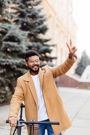 Young indian man in coat showing peace gesture with vintage bicycle on the city streetの写真素材