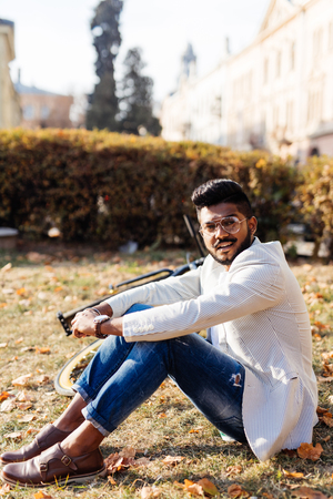Portrait of bearded indian man resting at park, sitting on grass near the bicycle, looking at camera. Leisure concept.の写真素材