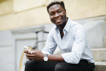 Handsome young Afro American man in casual wear is using smartphone while sitting on stairsの写真素材