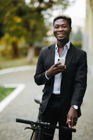 Handsome Afro American man in casual clothes is using smartphone, looking at camera and smiling while leaning on his bike, standing outdoorsの写真素材