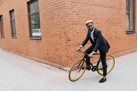 Stylish confident african businessman ride with his bicycle at building brick wall, copy spaceの写真素材