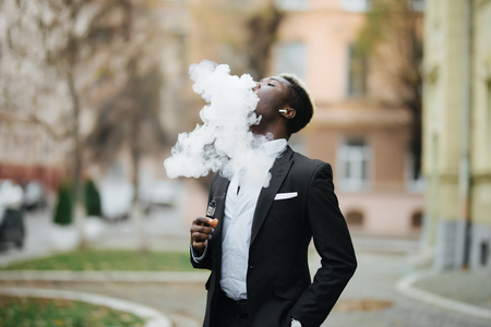 Handsome afro american business man with e-cigarette with smoke cloude standing on the city streetの写真素材