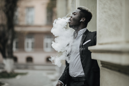 Handsome afro american business man with e-cigarette with smoke cloude standing on the city streetの写真素材