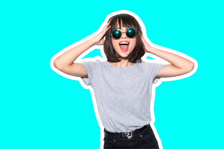 Collage in magazine style with colorful fashion girl in t-shirt and sunglasses scream holding her head toned blue backgroundの写真素材