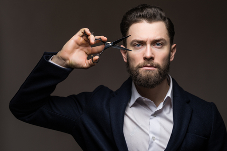 Barbershop advertising concept. Businessman with serious face isolated on black background. Man with long beard holds steel scissors. Macho in formal suit cuts beard and moustache.の写真素材