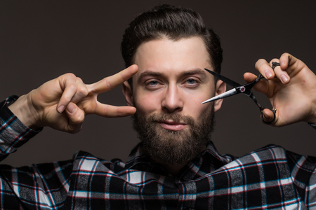 Bearded man, young man in plaid shirt holding Barber scissors and straight razor, near his face isolated on dark background.の写真素材