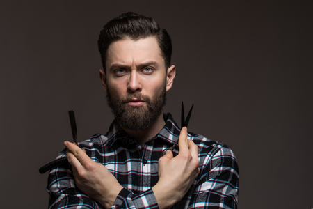 Bearded man, young man in plaid shirt holding Barber scissors and straight razor, near his face isolated on dark background.の写真素材