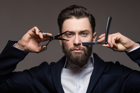 Young bearded man holding Barber scissors and razor near his face standing isolated on gray backgroundの写真素材