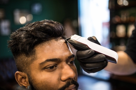 Unshaven man being clipped with professional electric shearer machine in barbershop. Male beauty treatment concept. Indian guy trim beard and mustache at barber shop salonの写真素材