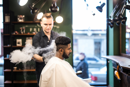 Bearded man with long beard, brutal, indian hipster with moustache, with stylish hair, haircut, getting powder on skin with make up brush by barber or hairdresser hands at barbershop