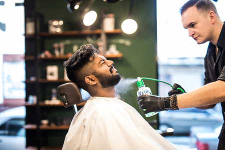 Smiling bearded barber combing and spraying hair of little kidの写真素材