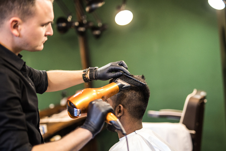 Barber with hairdryer drying and styling hair of client. Barber with hairdryer works on hairstyle for bearded man, barbershop background. Hipster bearded client getting hairstyle.の写真素材