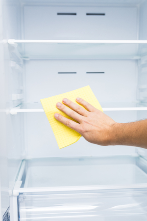 man's hand in yellow rubber protective glove cleaning white open empty refrigerator with green ragの写真素材