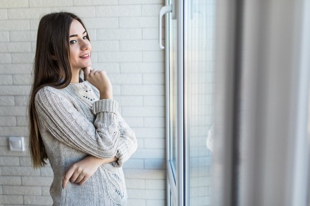 Portrait of a thoughtful attractive female looking outside through a window of an hotel room or homeの写真素材