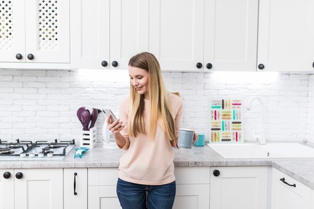 Young woman using smartphone leaning at kitchen table with coffee mug and organizer in a modern home. Smiling woman reading phone message. Brunette happy girl typing a text messageの写真素材
