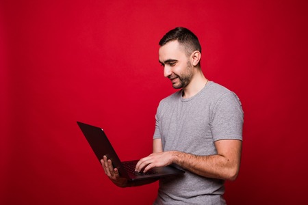 Young bearded man smiling while holding and using laptop over red backgroundの写真素材
