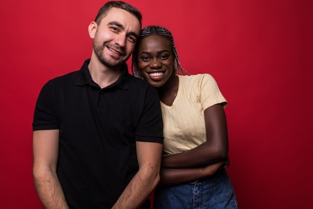 Portrait of smiling young interracial couple hugging and looking at camera isolated on the red backgroundの写真素材