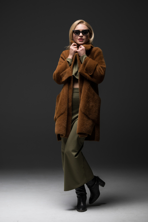 Smiling young woman with blonde hair wearing a coat and sunglasses over gray background.の写真素材