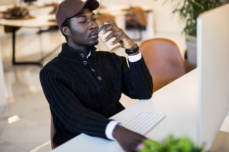 Image of young African businessman with cup of coffee looking at laptop screen at workplaceの写真素材