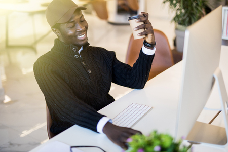 Image of young African businessman with cup of coffee looking at laptop screen at workplaceの写真素材