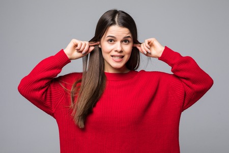 Portrait of disgusted young woman in black fur sweater covering ears with fingers on white wall backgroundの写真素材