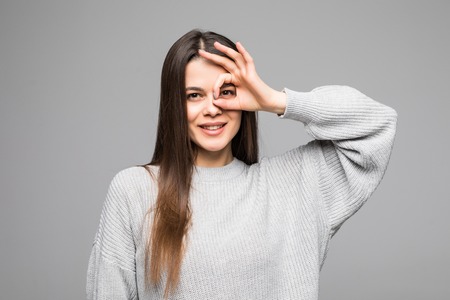 Close up portrait of beautiful joyful woman smiling, demonstrating white teeth, looking at the camera through fingers in okay gesture.の写真素材