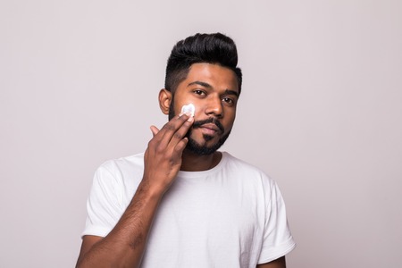 Portrait satisfied young indian man applying facial cream isolated over white backgroundの写真素材